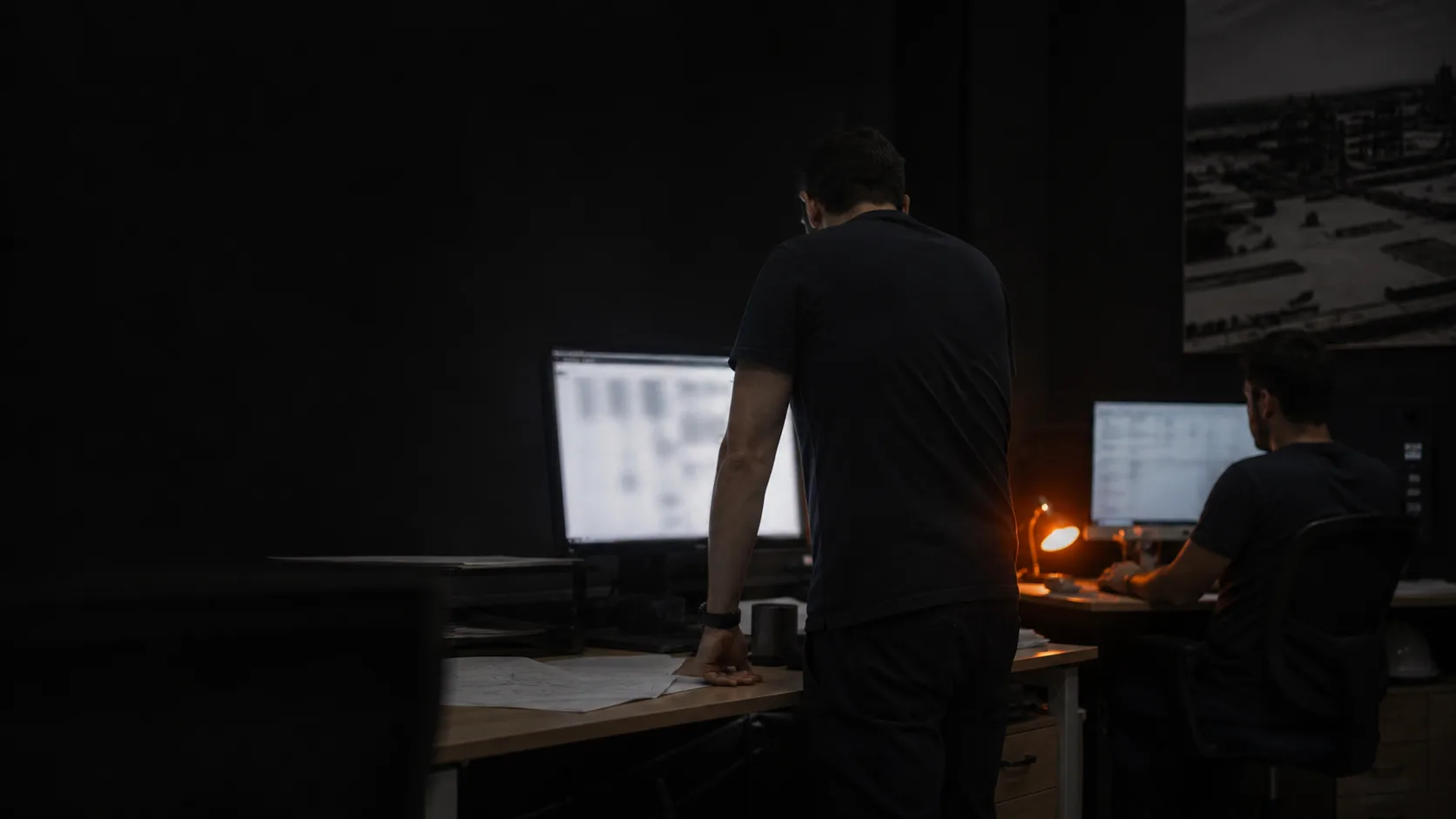 Dois profissionais trabalhando à noite num escritório escuro, iluminados pelas telas dos monitores e por uma luminária de mesa