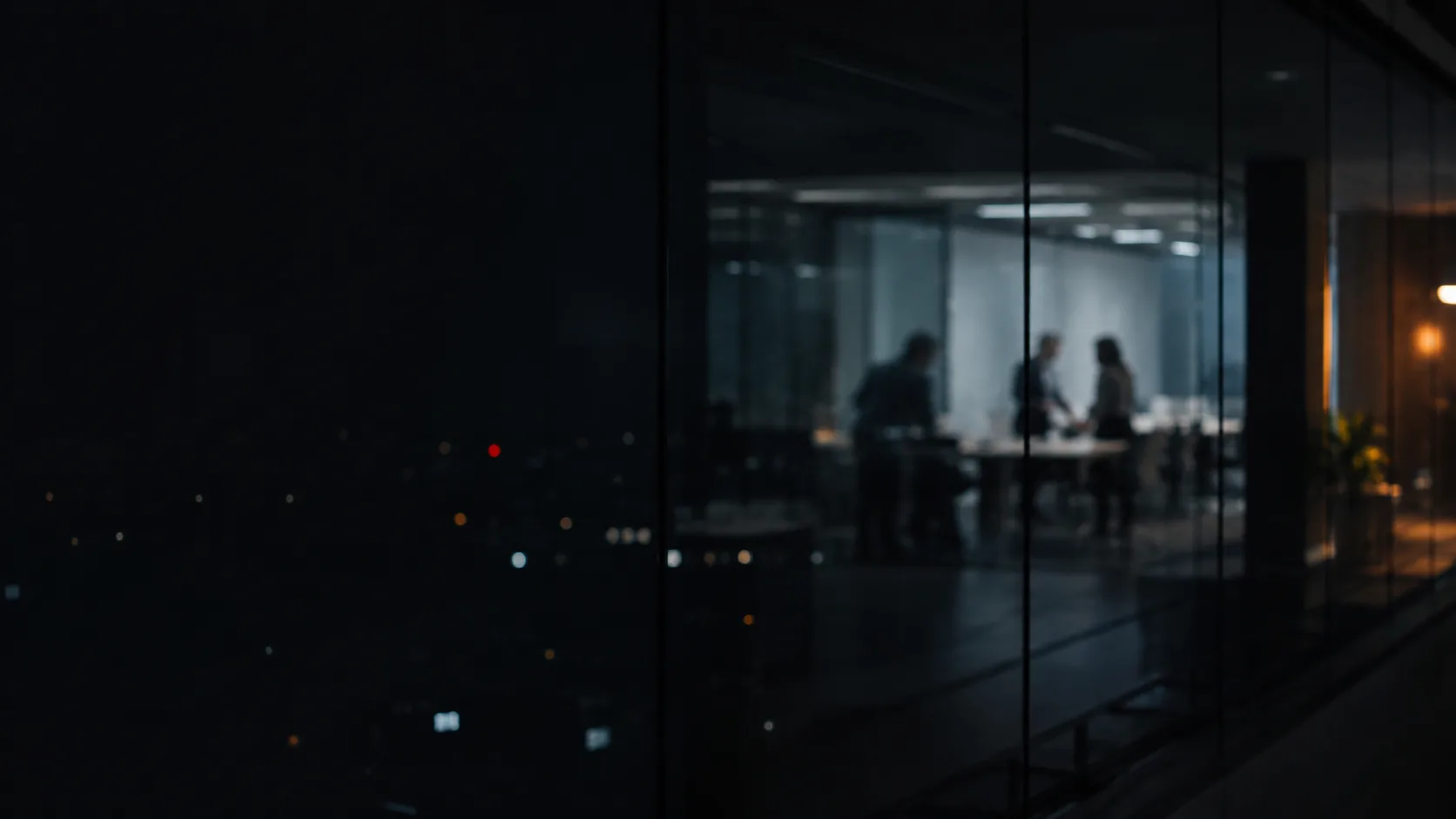 Sala de reunião vista de fora através de uma parede de vidro escura à noite, com três silhuetas desfocadas conversando ao redor de uma mesa