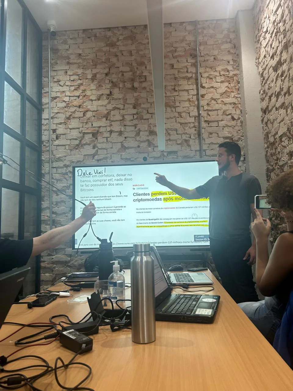Homem apontando para uma tela com um slide, numa sala de parede de tijolo aparente