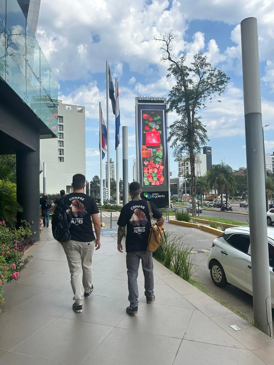 Dois integrantes da DSEC caminhando numa avenida, com bandeiras e prédios de vidro ao fundo