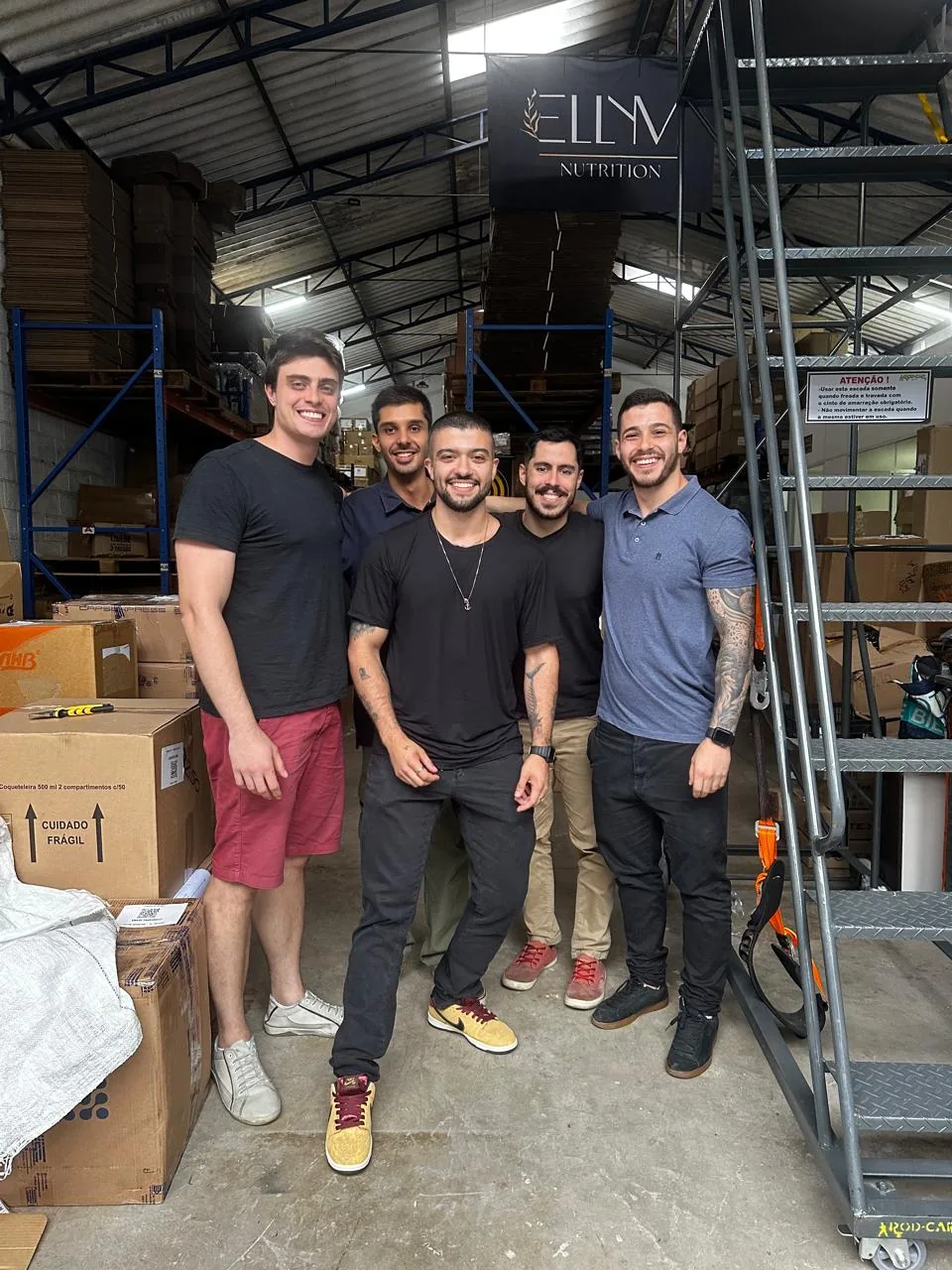 Cinco pessoas posando num galpao de estoque, entre prateleiras e caixas