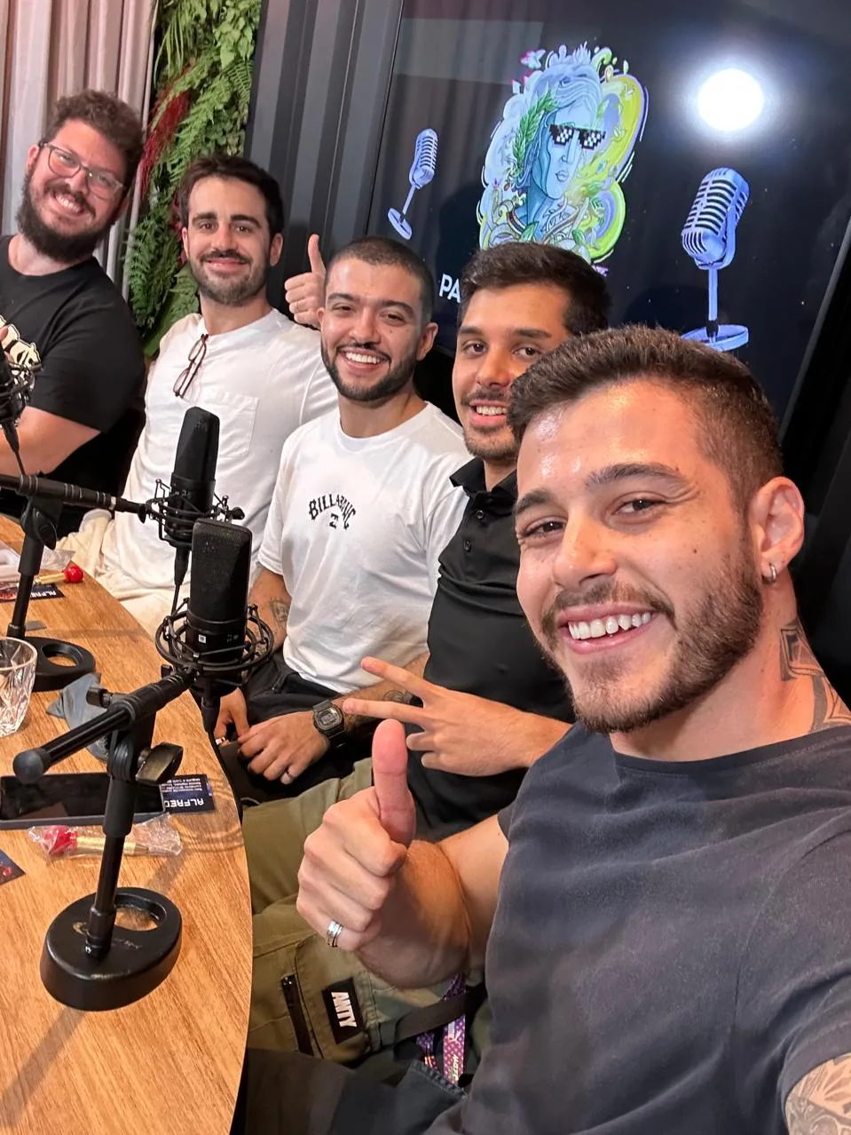 Cinco pessoas num estudio de podcast, microfones na mesa, uma delas faz selfie em primeiro plano