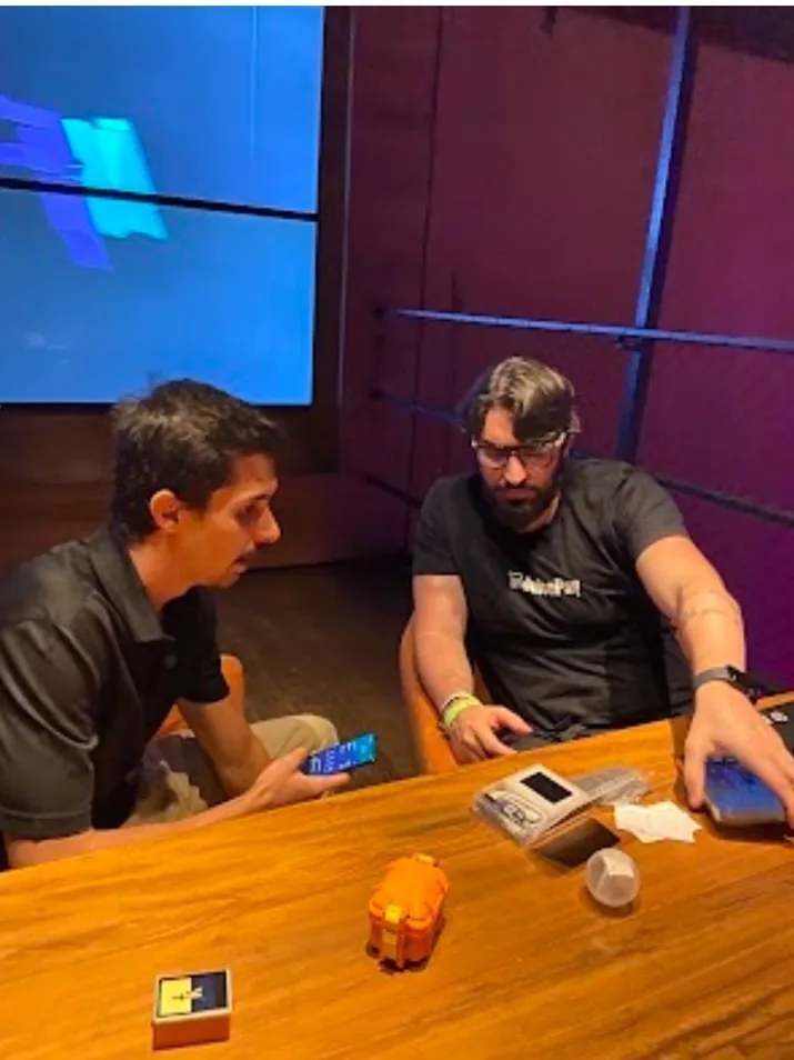 Dois homens sentados a uma mesa com um COLDKIT laranja e uma hardware wallet entre eles